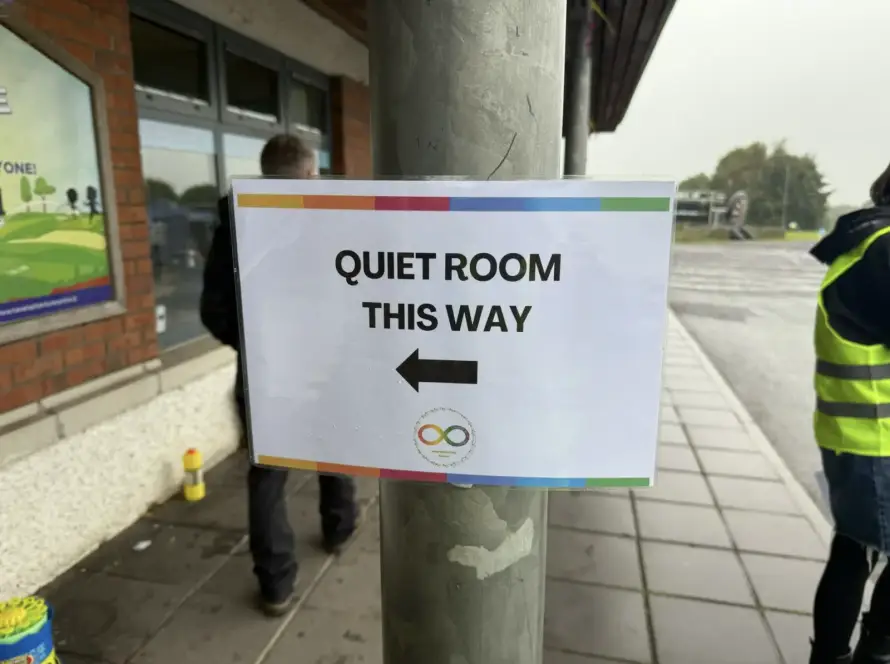 A sign on a pole reads "QUIET ROOM THIS WAY" with a left-pointing arrow. The sign is outdoors near a building, and a few people are visible in the background.