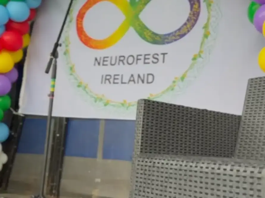 A stage decorated with colourful balloon arches, a microphone, and a wicker chair. A large sign reads "NEUROFEST IRELAND" with an infinity symbol in rainbow colours featuring human figures.