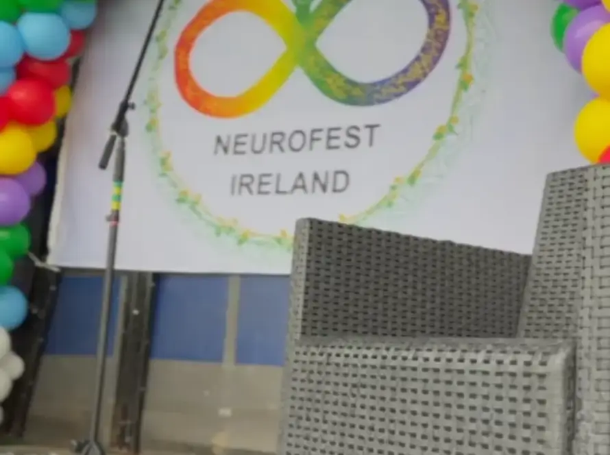 A stage decorated with a colourful balloon arch, a microphone stand, and a wicker chair. A banner reads "Neurofest Ireland" with an infinity symbol and two figures in rainbow colours.